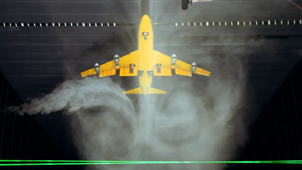 A yellow model airliner passing through a sheet of smoke in a test hall, its wingtip vortices swirling below