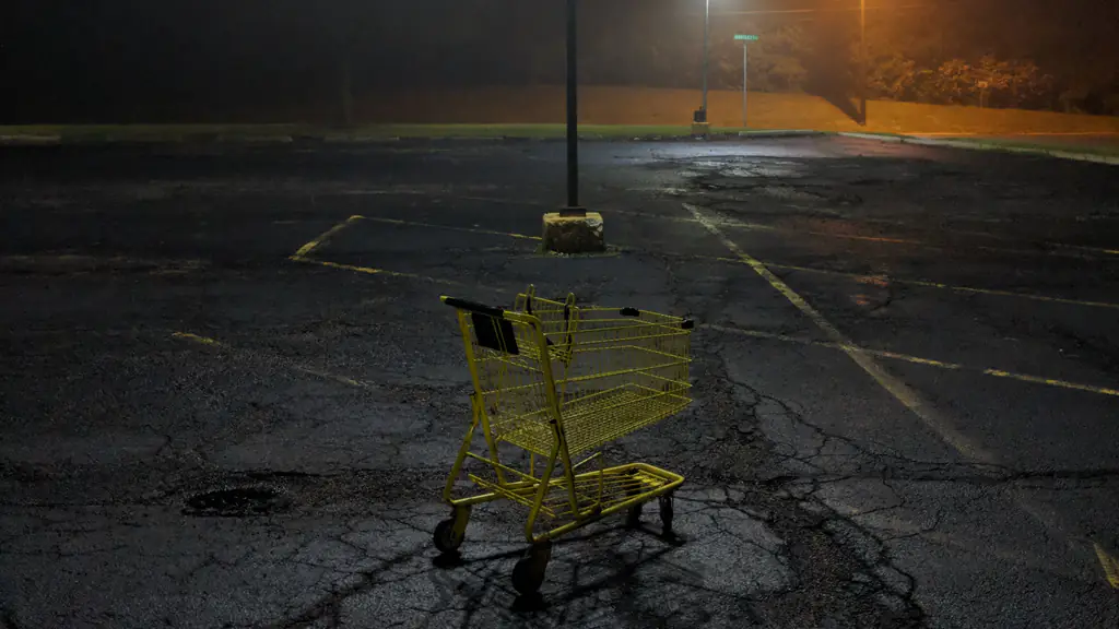 An empty yellow shopping cart stands alone in a cracked, deserted car park at night under a street lamp